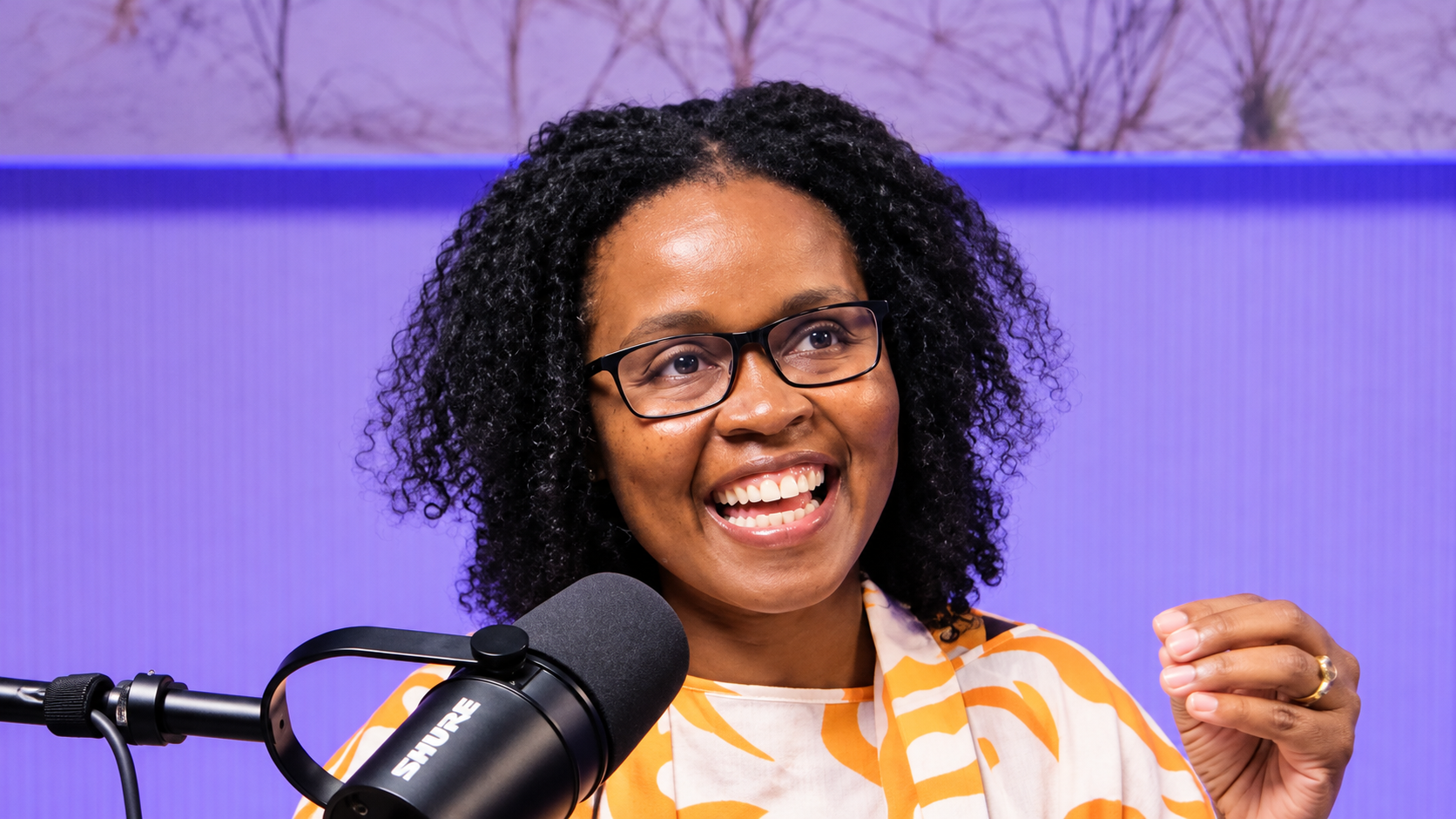 Woman in an orange and white patterned top speaking into a black Shure microphone against a purple background